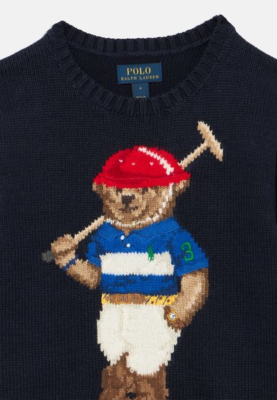 Granatowy sweter dziany z wizerunkiem misia w niebiesko-pasiastym polo i białych szortach, z czerwoną czapką, trzymającego kij do golfa. Obecne logo Ralph Lauren.