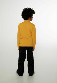 Gelbes Langarmoberteil mit hohem Kragen, kombiniert mit schwarzen, locker sitzenden Hosen. Der Stoff wirkt glatt mit minimalen Details.