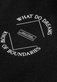 Sort stof med hvid broderet tekst, med sætningerne "WHAT DO DREAMS" og "KNOW OF BOUNDARIES," samt et logo i midten.