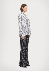 Λευκό πουκάμισο με μακριά μανίκια με σχέδιο μαύρης αλυσίδας και κείμενο. Συνδυασμένο με μαύρα φλορά παντελόνια που διαθέτουν παρόμοιο σχέδιο και υφή.