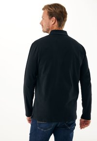 Maglietta nera a maniche lunghe con colletto, caratterizzata da una texture liscia e un taglio dritto. Abbinata a jeans blu scuro.