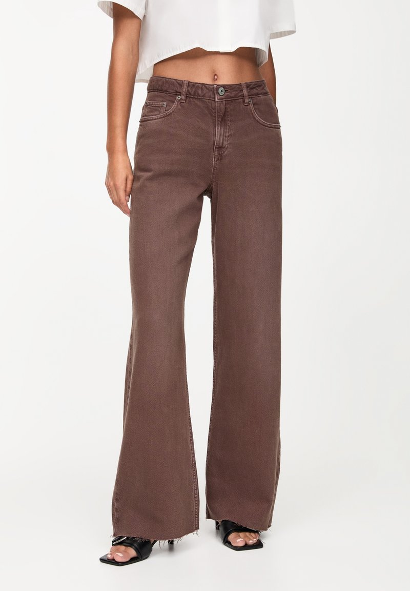 Jean évasé marron en denim, avec une taille mi-haute, des poches avant, et des finitions à cru au niveau des chevilles.