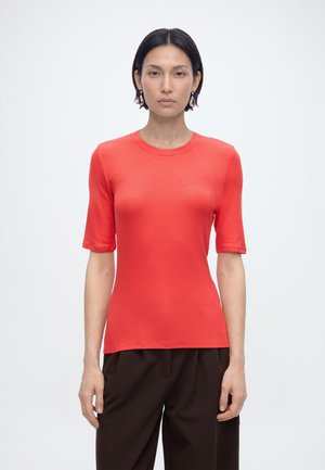 Kobieta o krótkich czarnych włosach ubrana w jasnoczerwony krótki rękaw i ciemnobrązowe spodnie, stojąca na jednolitym białym tle.