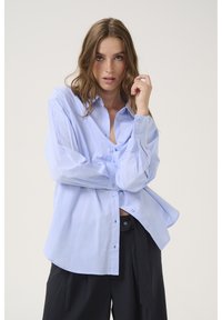 Light blue striped oversized shirt with button-up front and cuffed sleeves, made of a smooth fabric. Paired with dark pinstriped pants.