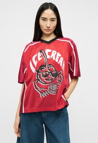 Νεαρή γυναίκα με μακριά μαύρα μαλλιά φορά χαλαρό κόκκινο αθλητικό μπλουζάκι με σκίτσο χαρακτήρα παγωτού και λευκό κείμενο "ICE CREAM", συνδυασμένο με μπλε τζιν παντελόνι.