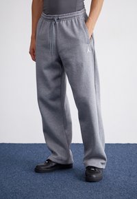 Szare spodnie dresowe z elastycznym paskiem i sznurkiem, z boczną kieszenią oraz białym logo na lewym udzie. Noszone z czarnymi butami.