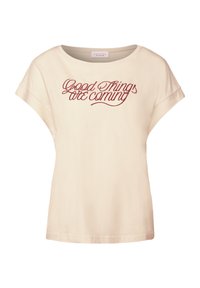 Camiseta de algodón beige con cuello redondo y mangas cortas enrolladas. Presenta texto en cursiva roja: "Cosas buenas están por venir." Textura suave.