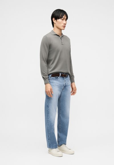 Άνδρας που φορά γκρι πόλο με μακριά μανίκια, ανοιχτόχρωμα μπλε τζιν με καφέ ζώνη και λευκά αθλητικά παπούτσια, στέκεται μπροστά σε απλό λευκό φόντο.