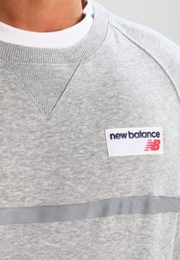 Felpa grigia chiara con collo a costine e texture. Presenta un'etichetta bianca con il logo "new balance" e una striscia orizzontale grigia.