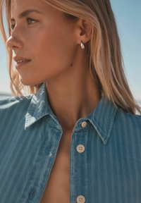 Chemise en denim bleu clair avec des rayures verticales, dotée d'un col classique et d'une fermeture à boutons. De simples boucles d'oreilles en argent en forme de goutte complètent le look.