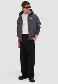 Joven vestido con gafas de sol negras, sudadera con capucha gris oscuro y cremallera, camiseta blanca con logo, pantalones negros de pierna ancha y zapatos negros gruesos, de pie con la mano en el bolsillo.