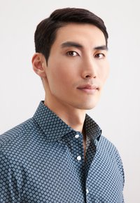 Camicia blu scuro con motivo geometrico, caratterizzata da accenti circolari bianchi. La camicia ha un colletto a punta e un'apertura con bottoni.