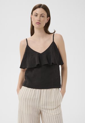 Femme portant un top noir sans manches à volants et un pantalon beige taille haute avec de fines rayures verticales, debout les mains dans les poches.