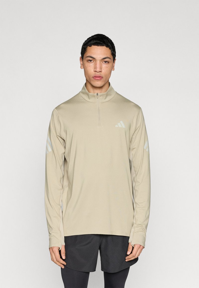 Μπεζ αθλητικό τοπ με μακριά μανίκια και φερμουάρ στο λαιμό, με αντανακλαστικές τρεις ρίγες στα μανίκια και λεία, ελαφριά ύλη.