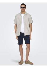 Beige linshort-sleeved skjorte, hvit t-skjorte under, marineblå shorts og beige sandaler. Modellen har på seg store solbriller.