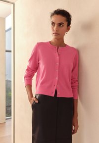 Lyserød cardigan lavet af blødt stof med rund halsudskæring, knaplukning og ribbede manchetter. Kombineret med en sort nederdel.