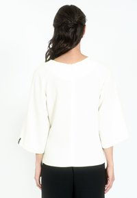 Blusa blanca con mangas acampanadas de 3/4, tela suave texturizada, cierre de cremallera en la parte posterior y corte recto. Llevada con pantalones negros.