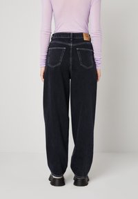 Osoba odjevena u crne široke traperice visokog struka s džepovima na stražnjoj strani, svijetlopurpurnu dugih rukava i crne debele cipele, stojeći okrenuta leđima.