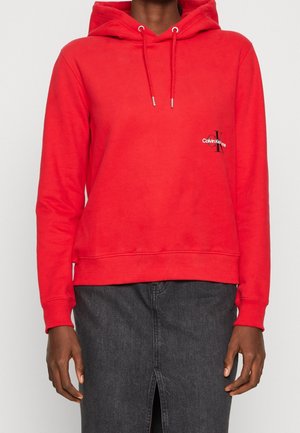 Personne portant un sweat à capuche rouge avec cordons et un petit logo Calvin Klein sur la poitrine, associée à une jupe en jean gris foncé avec une fente à l’avant.