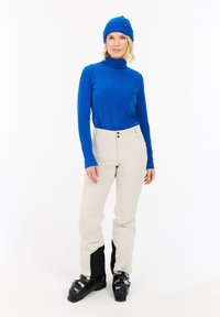 Blau gerippter Rollkragenpullover, cremige beige Skihosen mit zwei Knöpfen, schwarze Akzente an den Beinen, schwarze Skistiefel und eine passende blaue Strickmütze.