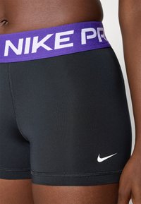 Mustat urheilushortsit, jotka on valmistettu venyvästä materiaalista. Niissä on violetti elastinen vyötärö, jossa lukee valkoisella tekstillä "NIKE PRO" sekä pieni valkoinen logo.