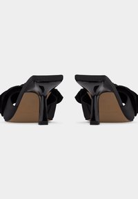 Μαύρες μπαλαρίνες mule με ψηλό τακούνι, τετράγωνα δάχτυλα και χοντρά τακούνια, εμφανιζόμενες από πίσω σε λευκό φόντο.