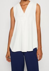 Haut blanc sans manches avec un col en V et un tissu lisse et léger ; présente une silhouette fluide et des fentes latérales discrètes à l'ourlet.