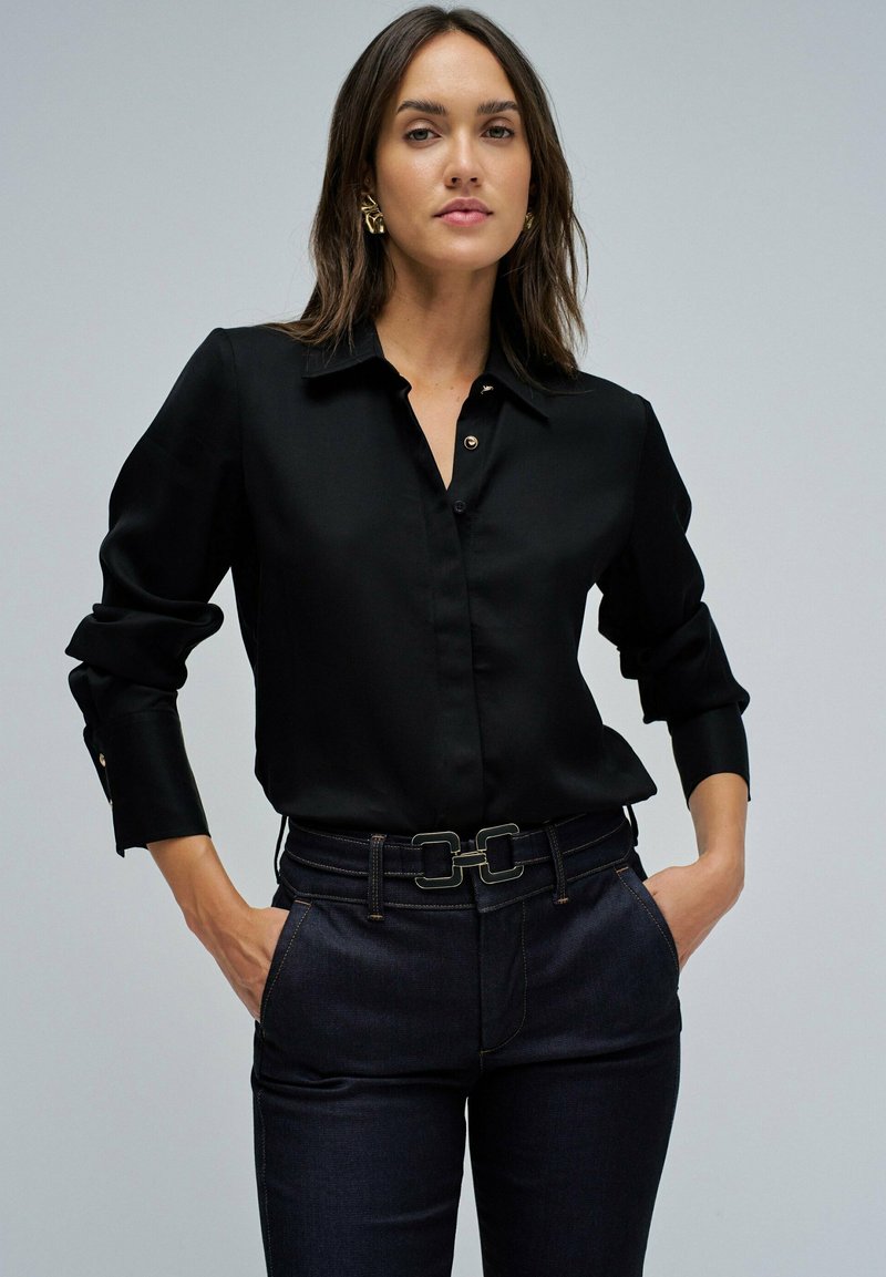 Chemise noire à manches longues avec col, accentuée de boutons dorés. Associée à un pantalon en denim foncé avec une ceinture à double boucle.