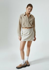 Beige overhemd met knopen, relaxed fit en lange mouwen; crèmekleurige wikkelrok; bruine loafers met dikke zwarte zolen; witte enkelsokken.