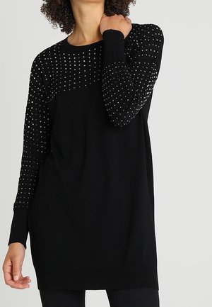 Robe-pull noire à manches longues avec de petits clous argentés sur le haut de la poitrine et les manches, portée par une personne aux cheveux bouclés.