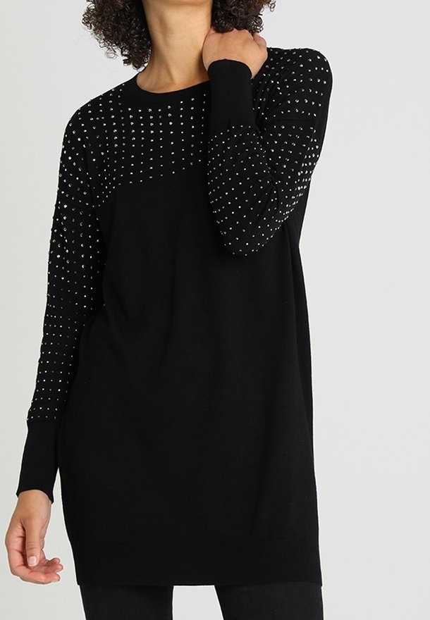 Robe-pull noire à manches longues avec de petits clous argentés sur le haut de la poitrine et les manches, portée par une personne aux cheveux bouclés.