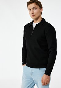 Zwarte geribbelde trui met een ritskraag, lange mouwen en een rechte zoom. Gerged over een wit shirt, gecombineerd met lichtblauwe denim.