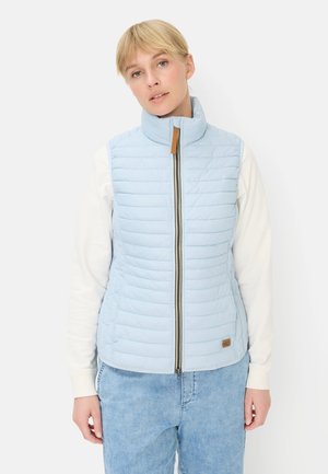 Femme aux cheveux blonds courts portant un gilet matelassé bleu clair sur une chemise blanche à manches longues et un jean bleu clair, sur fond blanc.