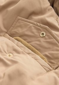 Beige quiltet stof med en trykknap-lomme, der har en tan fleece-foring, som fremhæver en glat tekstur og rene syninger.