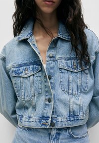 Blå denimjacka i cropped-stil med knappstängning framtill, två bröstfickor och en urtvättad look. Har en klassisk krage och metallknappar.