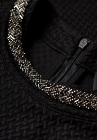 Vêtement en tissu noir texturé avec un col rond orné de petites perles argentées et une fermeture éclair noire visible à l'arrière.