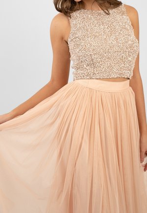 Kvinde iført ærmeløs beige crop-top med pailletter og matchende plisseret tulle-nederdel, holder skørtet ud til siden med den ene hånd.