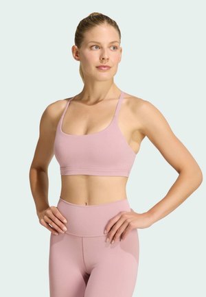Kobieta o blond włosach związanych w kucyk, ubrana w pasujący jasnoróżowy sportowy stanik i legginsy z wysokim stanem, stoi z rękami na biodrach i patrzy w prawo.