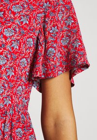Robe florale rouge avec des motifs complexes bleus et blancs. Manches courtes et amples avec un détail de taille froncée sur un tissu léger.
