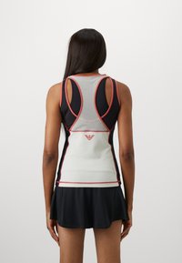 Top desportivo em preto e branco com inserções em malha e detalhes em vermelho. Apresenta uma saia rodada e um design de costas nadadora. Logótipo exibido nas costas.