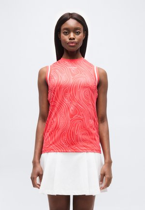 Naine, kes kannab punast varrukateta topi valgete keerutismustritega ja valget volditud seelikut, seisab ühevärvilise heleda tausta ees.