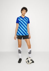 Blau- und türkisgestreiftes Sportshirt, schwarze Shorts, weiße Socken mit schwarzen Streifen und schwarze Fußballschuhe, mit einem Fußball zu den Füßen.