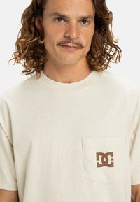 Beige bavlněné tričko s krátkým rukávem a kulatým výstřihem. Na levé straně hrudi má hnědý kapsový logo. Měkký, ležérní design.