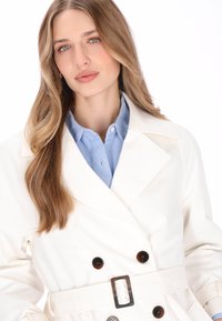 Hvid dobbeltradet trenchcoat med bælte i taljen, brune knapper som accenter og en glat stoftekstur over en lyseblå skjorte.