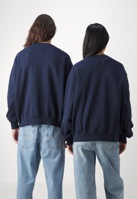 Sudaderas unisex de color azul marino con hombros caídos y puños acanalados, combinadas con jeans de mezclilla de color azul claro. La tela parece suave y texturada.