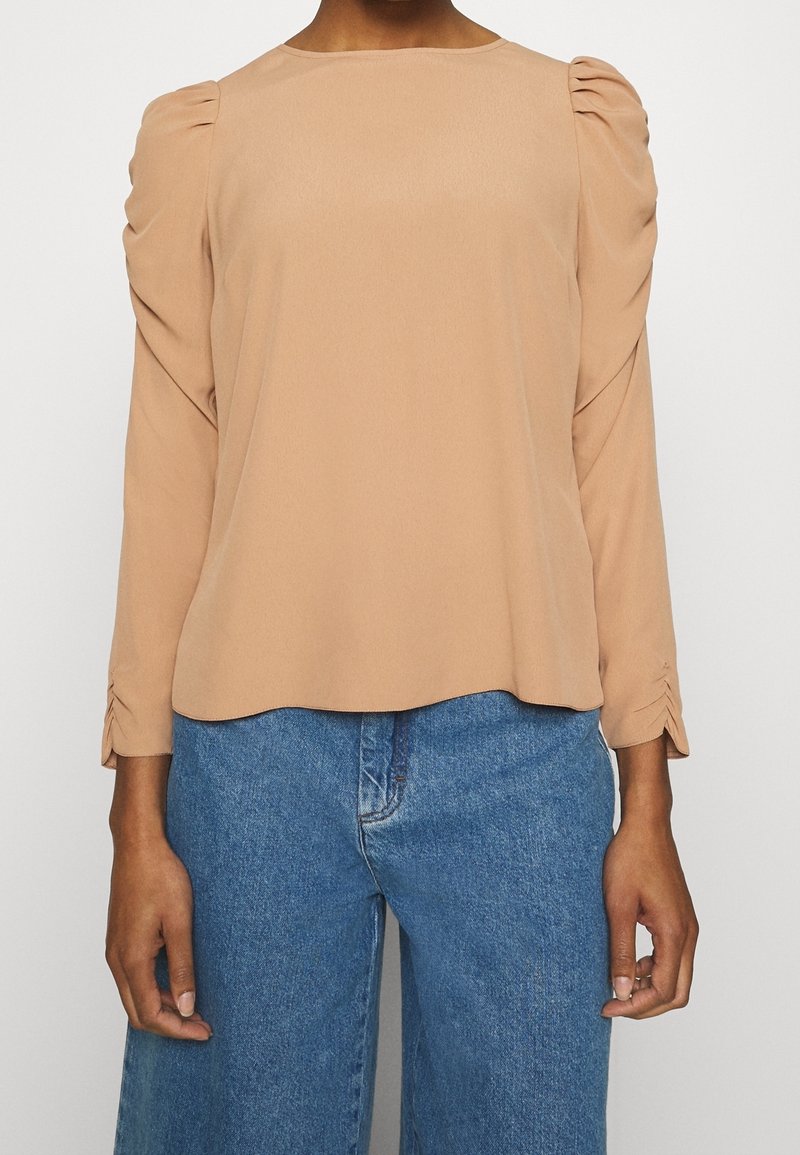 Blusa a maniche lunghe di un leggero color beige con spalle plissettate e polsini aderenti, abbinata a jeans a gamba larga blu. Tessuto dal tocco morbido.