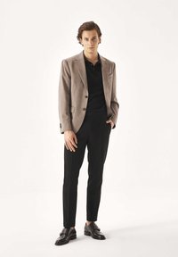 Grauer Blazer über einem schwarzen Poloshirt, kombiniert mit schwarzen Hosen und glänzenden schwarzen Schuhen. Schlanke Passform, glatte Textur und dezente Details am Blazer.