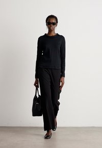 Zwart langemouwen top, zwarte wijde broek en zwarte ballerina's. Accessoires zijn een zwarte handtas en een donker zonnebril. Glad stofafwerking.