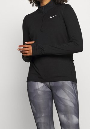Persoon draagt een zwart Nike-trainingsjack met lange mouwen en een ritssluiting, en grijze legging met patroon, staat met de handen voor zich gevouwen.