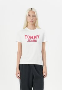 Camiseta de manga corta blanca con texto rojo "TOMMY JEANS". Hecha de algodón, con un corte clásico y escote redondo. Combinada con pantalones oscuros.